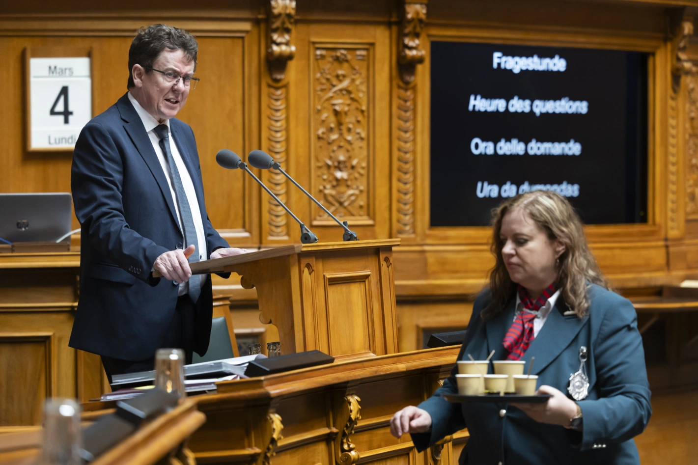 Bundesrat Albert Rösti spricht in der Fragestunde während Ratsweibelin Nathalie Radelfinger für die Verpflegung mit Kaffee sorgt. Foto: Peter Klaunzer (Keystone)